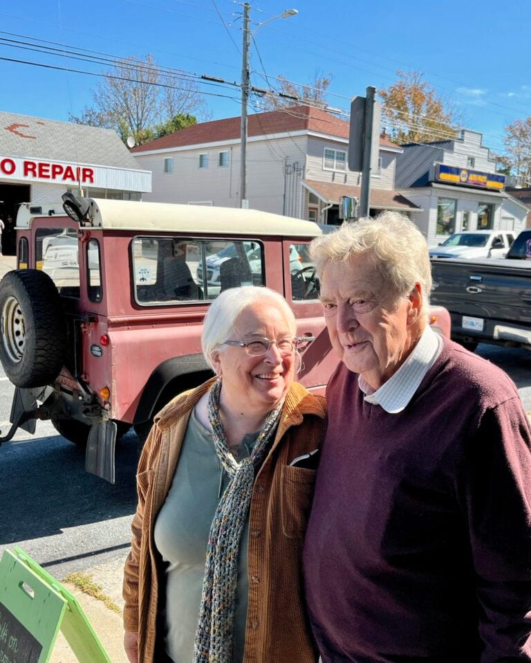 An older woman and man stand smiling together on a sunny street, with a red vehicle and buildings, including an auto repair shop, in the background.